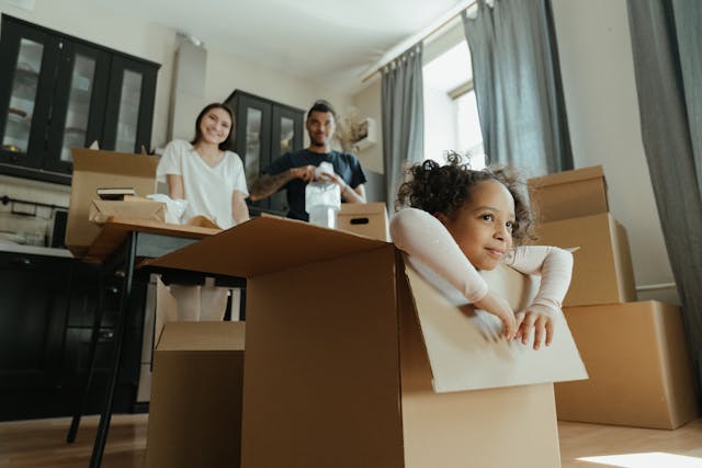 Child peering out from a large cardboard box while parents unpack in the background.